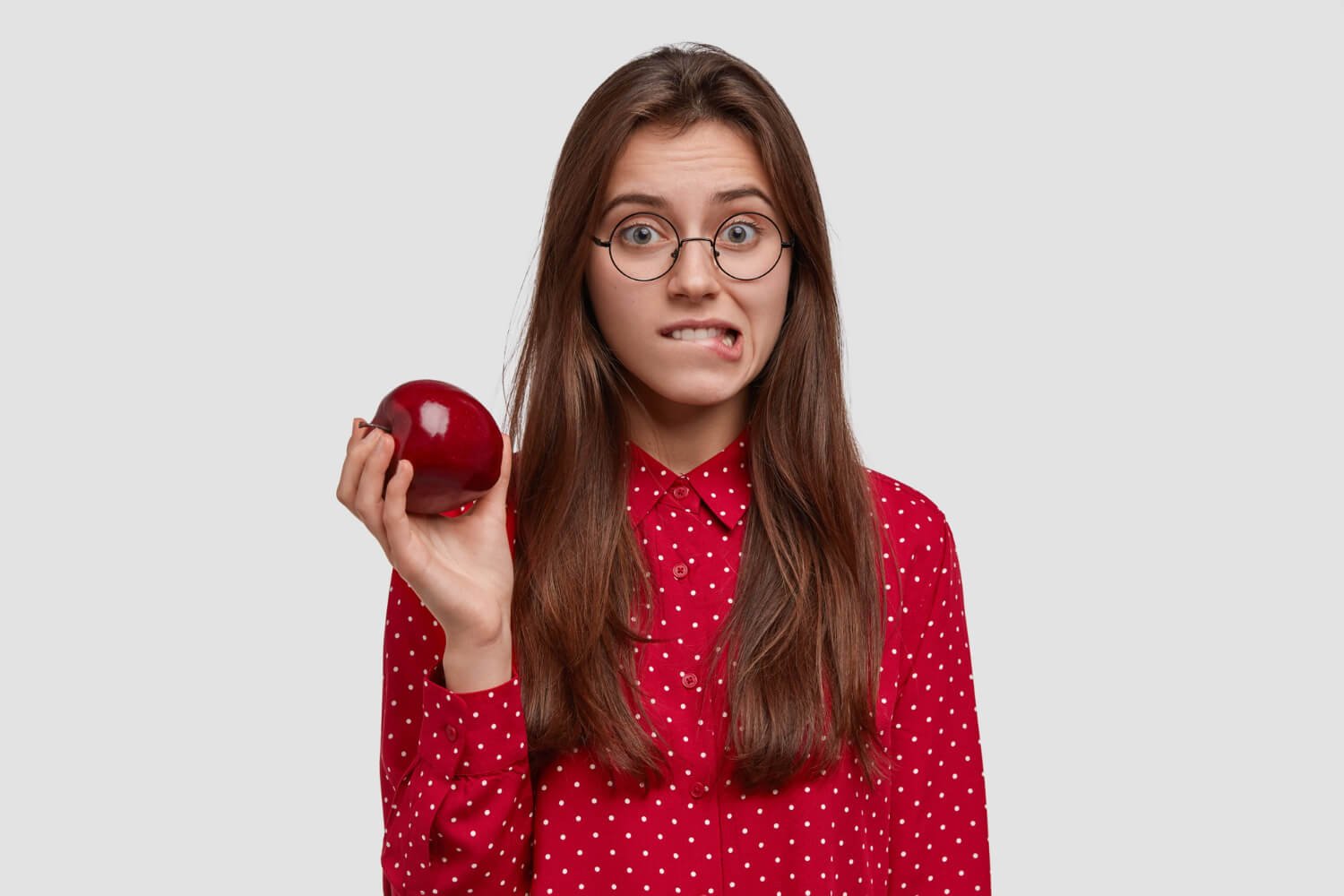 Mulher de óculos e camisa vermelha morde o lábio com dúvida ao segurar maçã, ilustrando cuidados com lentes de contato dental em São Bernardo do Campo.